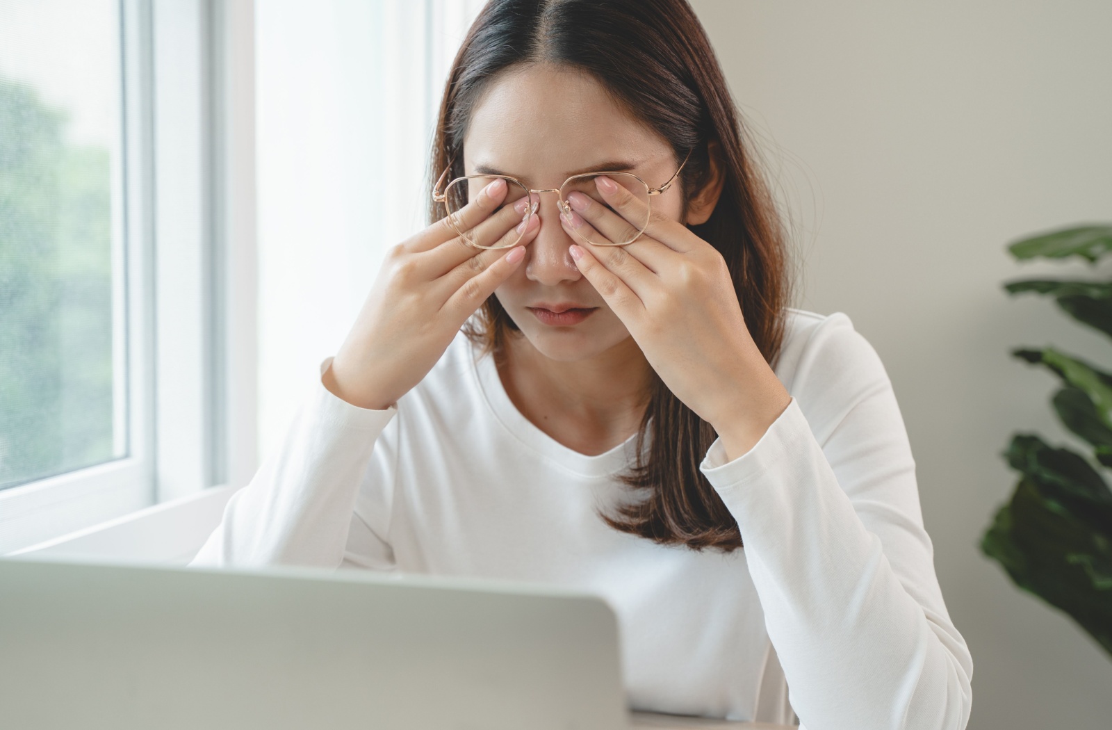 A woman working at a laptop rubs both eyes due to dry eye syndrome caused by MGD.
