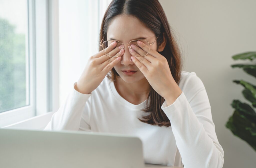 A young adult working at their computer, rubbing their eyes under their glasses due to dry eyes.