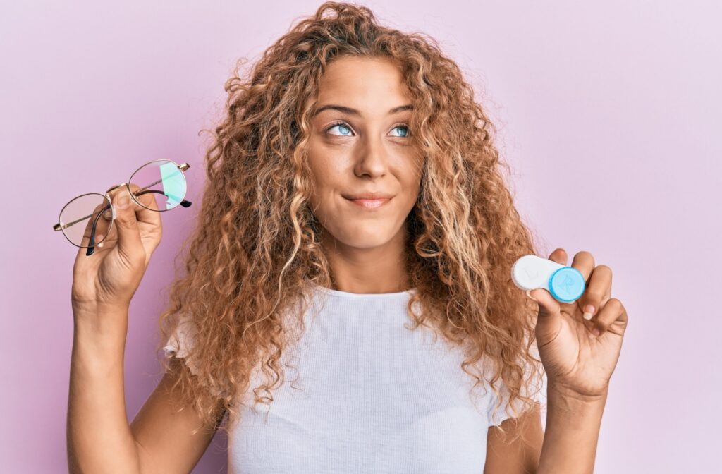 Against a pink background, a young adult with curly hair holds up a pair of glasses in one hand and contacts in the other