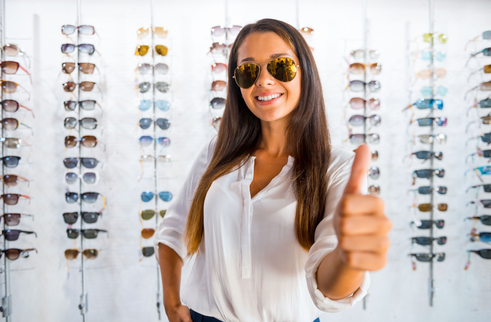 A patient giving a thumbs-up about their new sunglasses standing in front of an optometrist's display wall.
