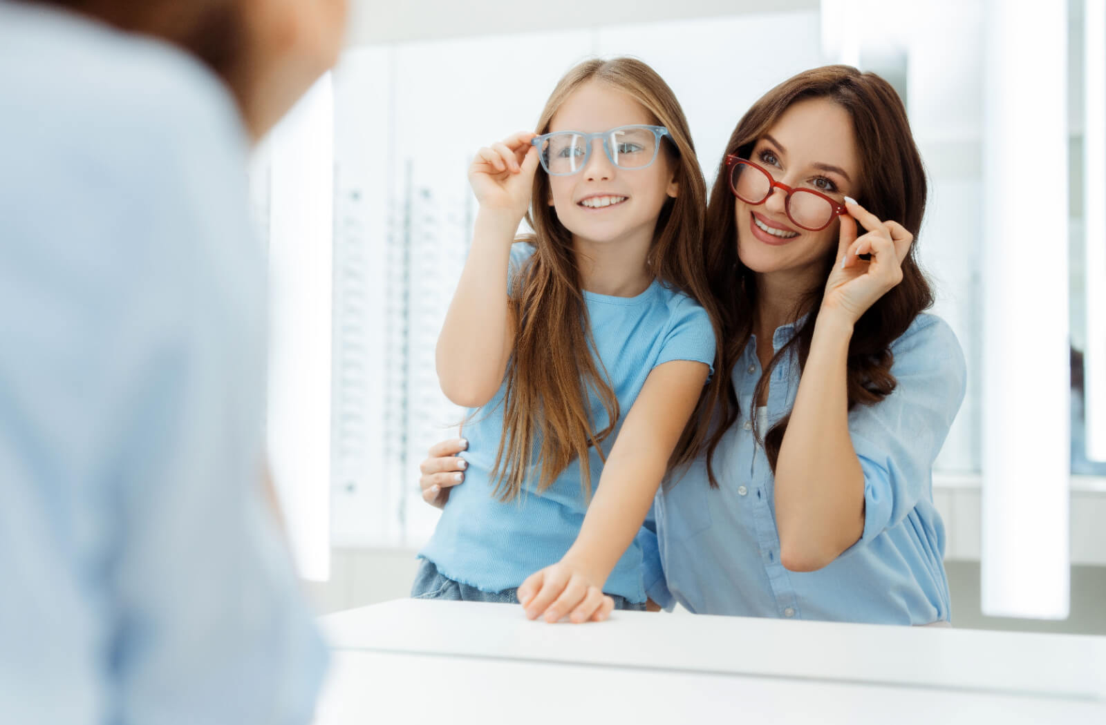 A patient and their young child smiling in the mirror while trying on a new pair of glasses.
