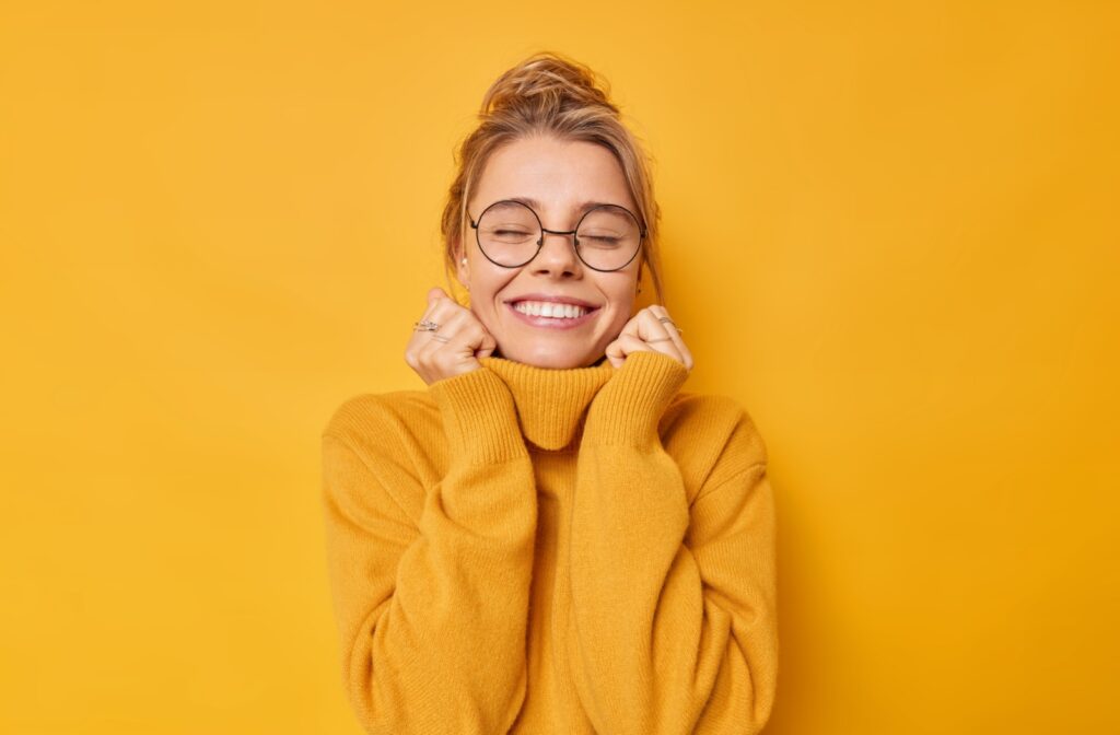 Against a yellow backdrop, a happy person wearing circular eyeglasses holds the collar of their yellow turtleneck sweater