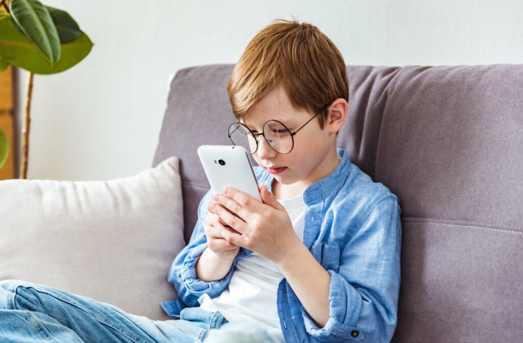 A young child with glasses sitting on the couch, holding a phone near their face due to nearsightedness.